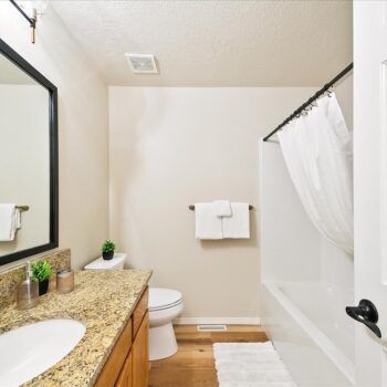 A modern bathroom featuring a speckled granite vanity, a framed mirror, a white toilet, and a bathtub with a white curtain.