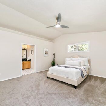 A bright, carpeted bedroom with a queen bed, a ceiling fan, and an open doorway leading to a bathroom.