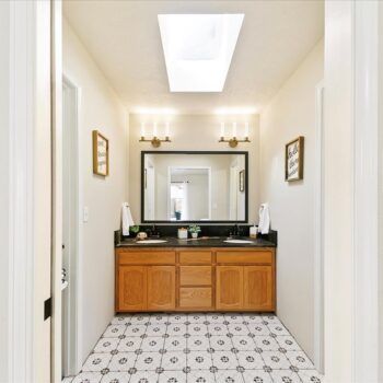 A bathroom with a double wood vanity, a large mirror, dual sinks, gold sconces, and patterned tile flooring.
