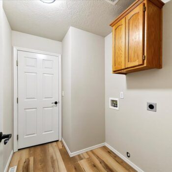 A laundry room showing a white door, light wood-style flooring, beige walls, and a wooden wall-mounted storage cabinet.