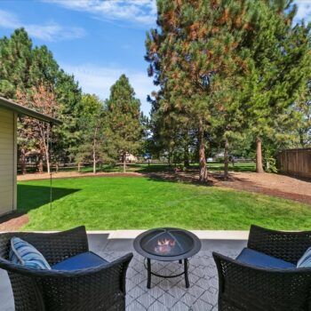 Two wicker armchairs with blue cushions sit on a concrete patio facing a fire pit, with a green lawn and trees beyond.