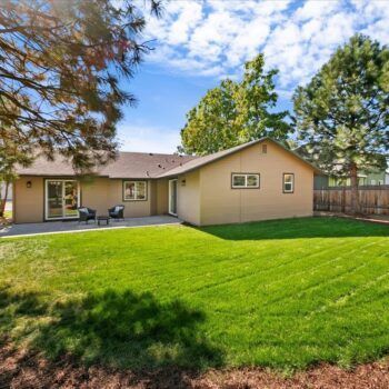 A tan, single-story house with a green lawn, concrete patio, and mature trees against a bright blue sky.