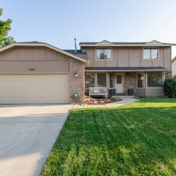 A two-story suburban home with tan siding, a brick accent wall, a two-car garage, and a lush green front lawn.