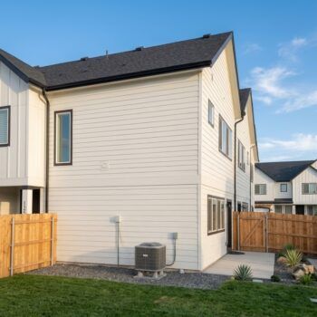 A side view of a modern two-story home with white horizontal siding, a dark roof, and a small backyard with a fence.
