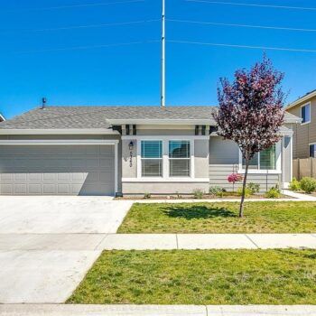 A one-story gray house with a two-car garage, white trim, and a decorative tree in the front yard under a blue sky.