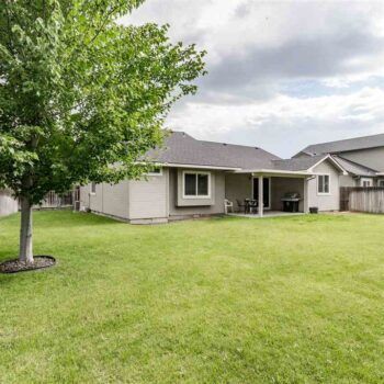 The back of a tan single-story house with a patio and a large grassy yard under a cloudy sky.