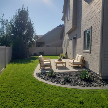 Patio set with wooden chairs and a table on a concrete slab next to a house, surrounded by grass and rock landscaping.
