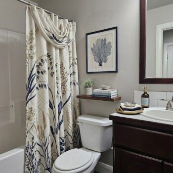 A bathroom with a nature-themed shower curtain, a white toilet, a floating wood shelf with books, and a dark vanity.