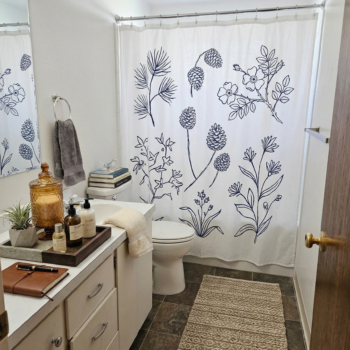 A bright bathroom featuring a white shower curtain with blue botanical line art, a wooden vanity, and a textured rug.