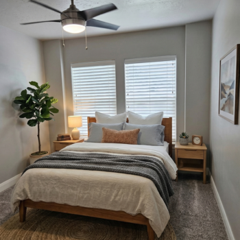 A neatly made bed with neutral bedding and a dark gray throw blanket in a bedroom with two windows and a ceiling fan.