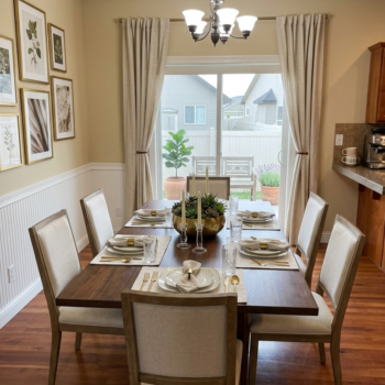 A dining room with a wooden table set for six, cream-colored chairs, beadboard wainscoting, and floor-length curtains.