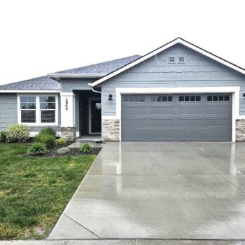 A single-story suburban house with blue siding, a stone facade, a gray garage door, and a wet concrete driveway.