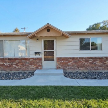 A single-story house with horizontal tan siding, a brick lower exterior, a central front door, and a concrete walkway.