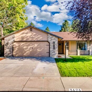 A beige ranch-style house with stone accents, a two-car garage, and a manicured green lawn under a partly cloudy blue sky.