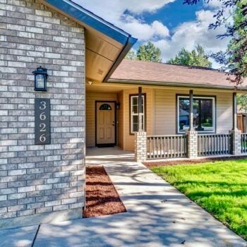 A house exterior featuring a grey brick wall with address 3626, a beige porch with railing, and a concrete walkway.