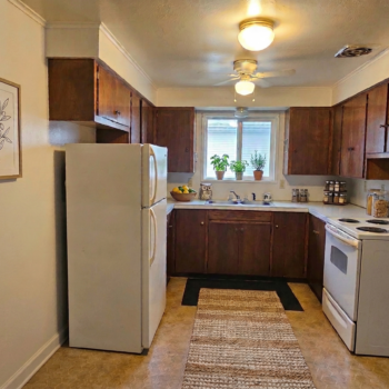 A view of a galley-style kitchen featuring wood cabinets, a white refrigerator, a white stove, and a window over the sink.