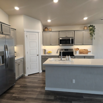 A modern kitchen with grey cabinets, stainless steel appliances, a white quartz island, and wood-look flooring.