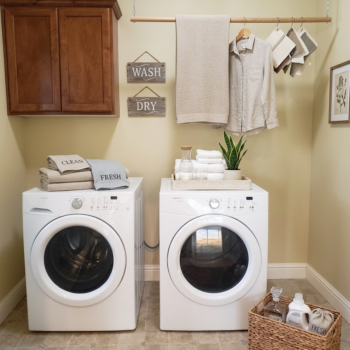 A bright laundry room with two white front-load machines, a wooden drying rack, folded towels, and a wicker basket.