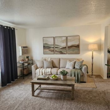 A neutral-toned living room with a beige sofa, three landscape art pieces, a coffee table with apples, and a floor lamp.