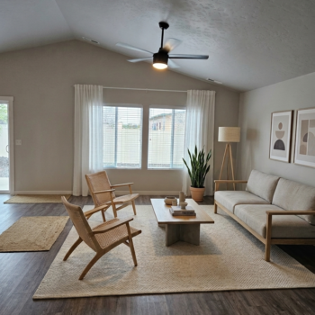 A modern living room with a neutral sofa, two wooden armchairs, a coffee table, and large windows with white curtains.
