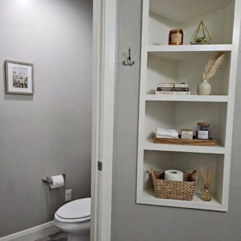A bathroom view with a white built-in shelving unit featuring home decor, a toilet, and a framed wall hanging.