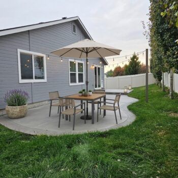 A dining set with an umbrella sits on a gray patio next to a house with gray siding, string lights, and a grassy lawn.