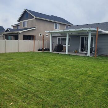 A residential backyard featuring a green lawn, a white vinyl fence, and a gray house with a white patio cover.