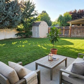 A patio with two chairs and a wooden coffee table, facing a grassy backyard with a garden shed and trees.