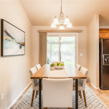 A modern dining room with a wooden table, white upholstered chairs, a geometric area rug, and a central chandelier.