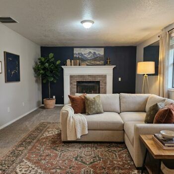 A cozy living room with a cream sectional sofa, dark blue accent wall, brick fireplace, and a patterned area rug.