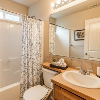 A bright bathroom with a light wood vanity, white sink, toilet, tub with a patterned shower curtain, and a large mirror.