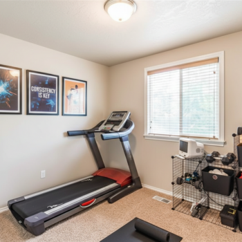 A home gym features a treadmill, a storage rack with weights and gear, three wall art frames, and a window.