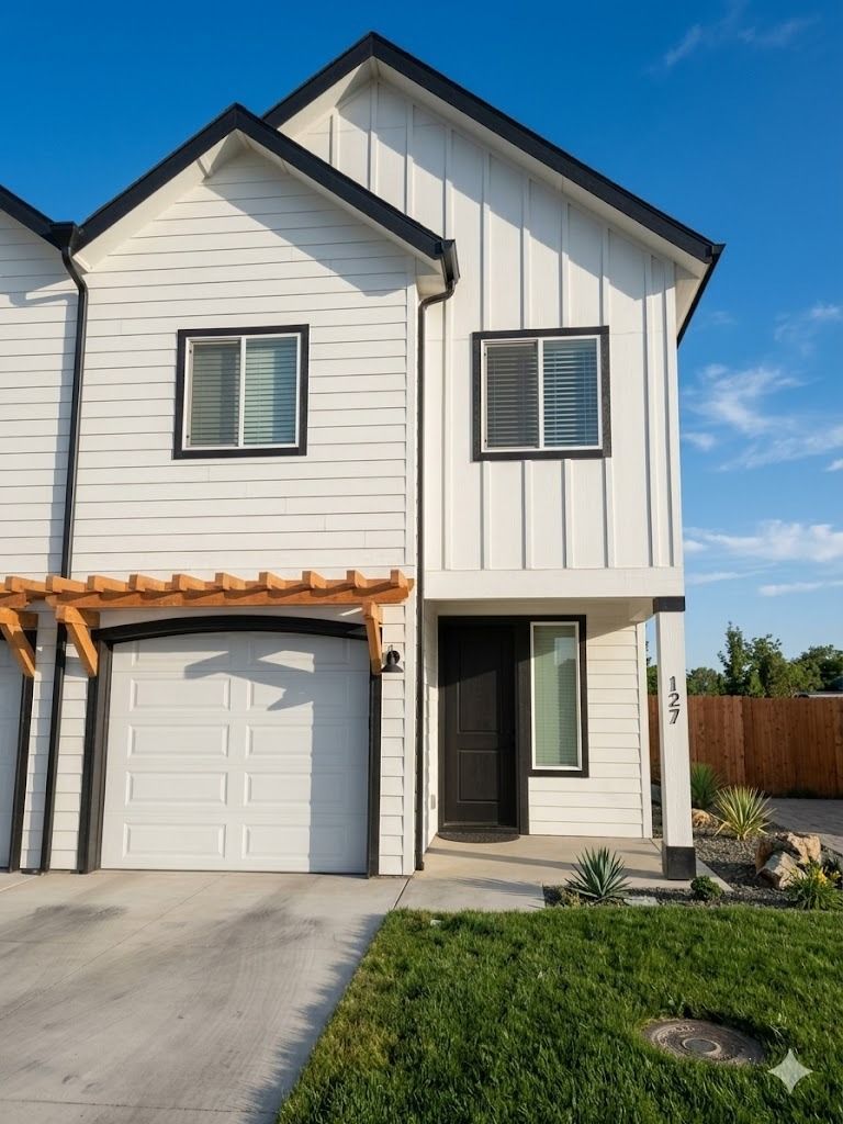 A white two-story townhouse with vertical siding, a single-car garage, and a wooden pergola under a clear blue sky.