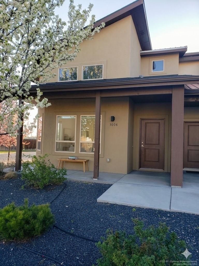 A modern tan two-story house with brown trim, a front porch, and a blooming tree in a landscaped yard.