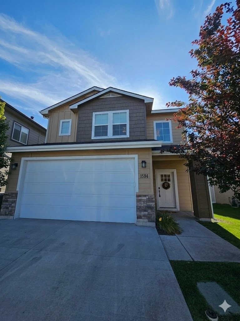 Two-story suburban house with a tan exterior, a white garage door, and brown stone accents under a sunny blue sky.