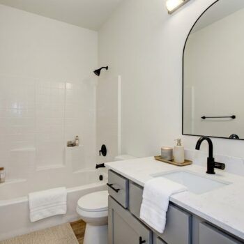 A bathroom with a white tub, toilet, grey vanity, white countertop, black faucet, and an arched mirror.