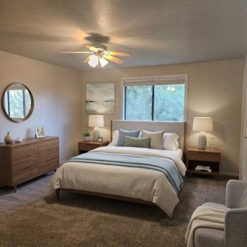 A bedroom with a bed, two nightstands, a dresser with a round mirror, and a ceiling fan.