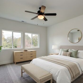 A modern, light-filled bedroom with a bed, bench, wooden dresser, and circular wall mirror under a ceiling fan.