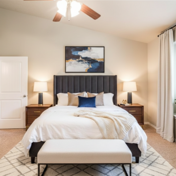 A modern bedroom with a dark upholstered headboard, white bedding, a bench, two bedside lamps, and abstract wall art.