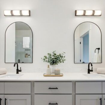A modern bathroom vanity with dual arched mirrors, black faucets, light gray cabinets, and two wall-mounted light fixtures.