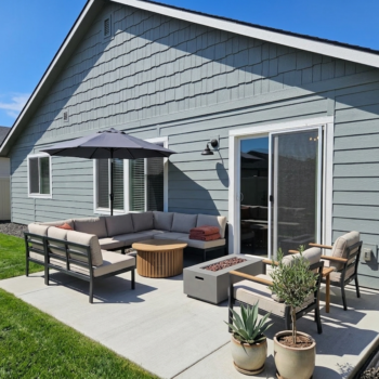 A patio set with a sectional sofa and fire pit on a concrete slab next to a light-blue-sided house on a sunny day.