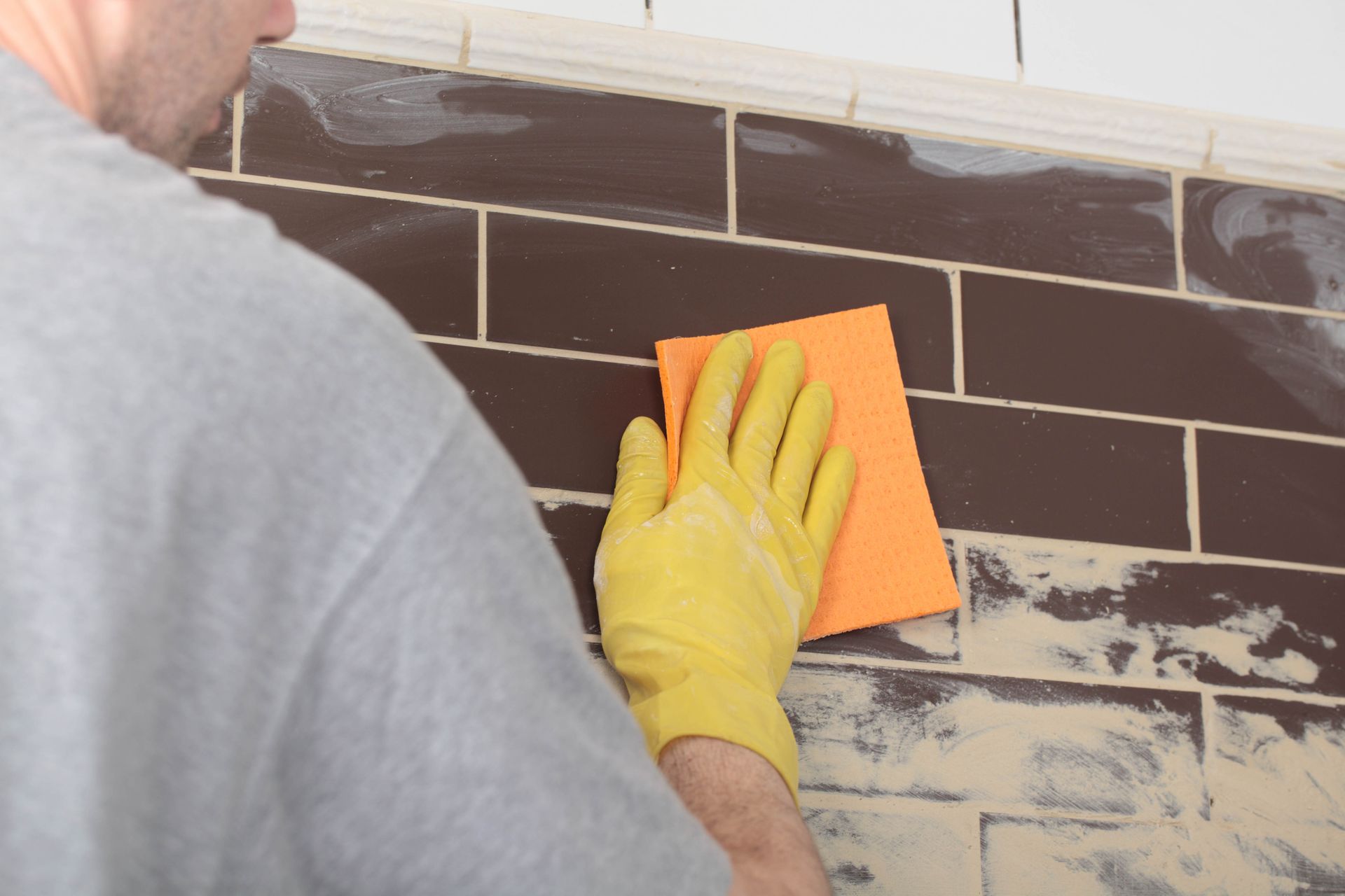 Person wearing yellow glove wiping brown tiled wall with orange sponge.
