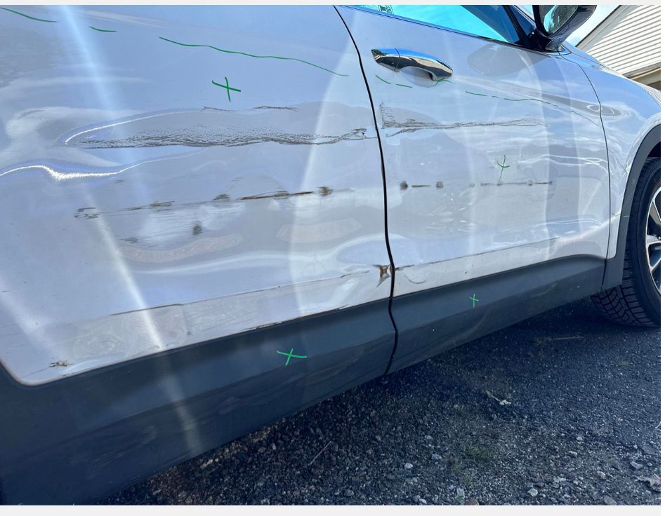 A white car with a damaged door is parked on the side of the road before