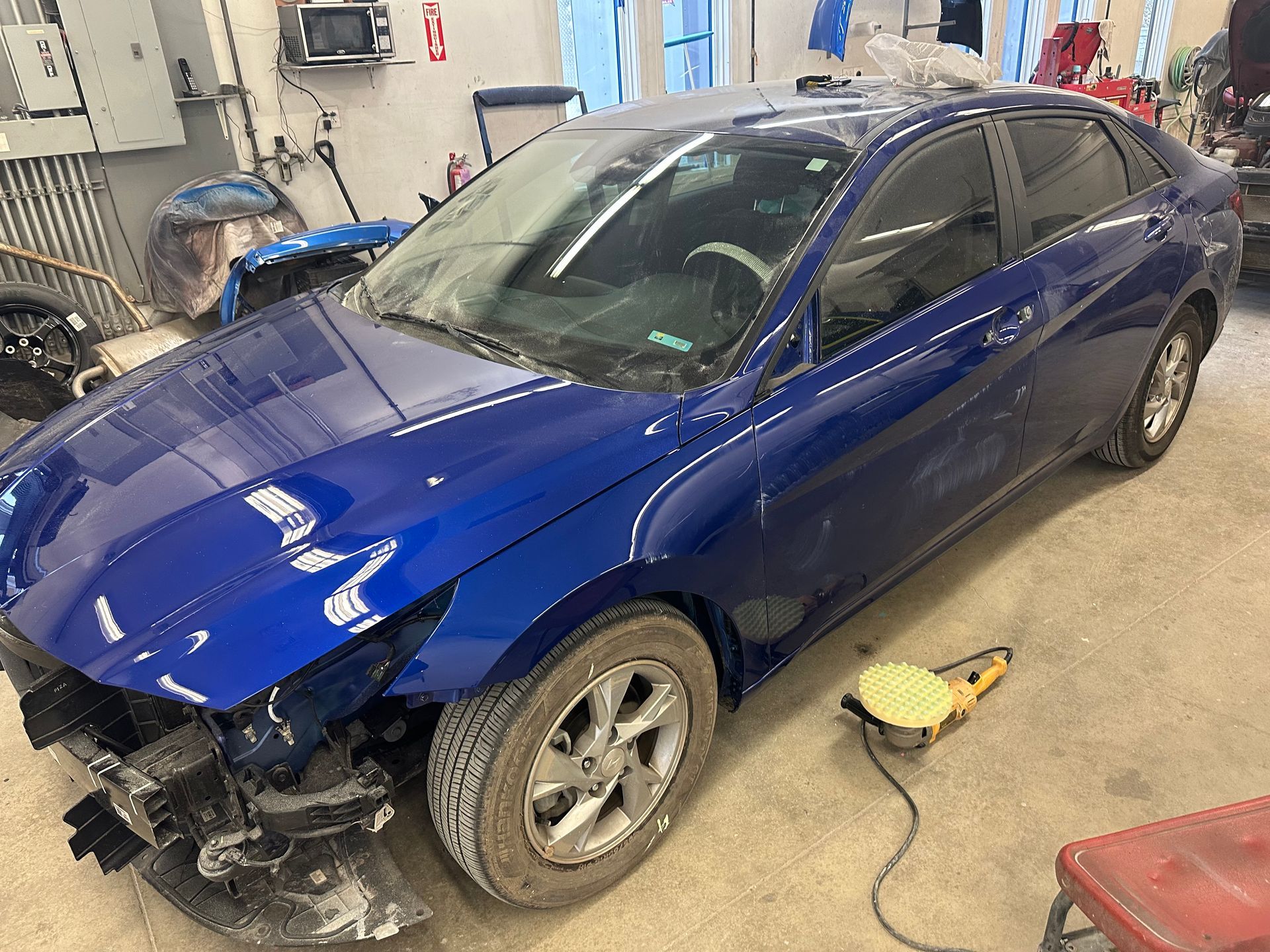 A blue car is sitting in a garage next to a yellow tool.