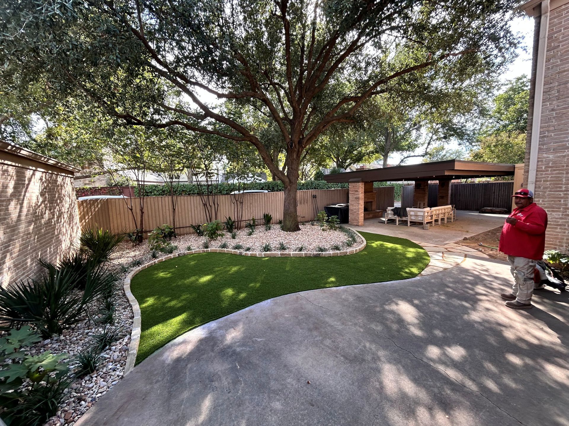 Backyard patio with large tree, curved lawn, stone border, and a person in a red jacket near the driveway