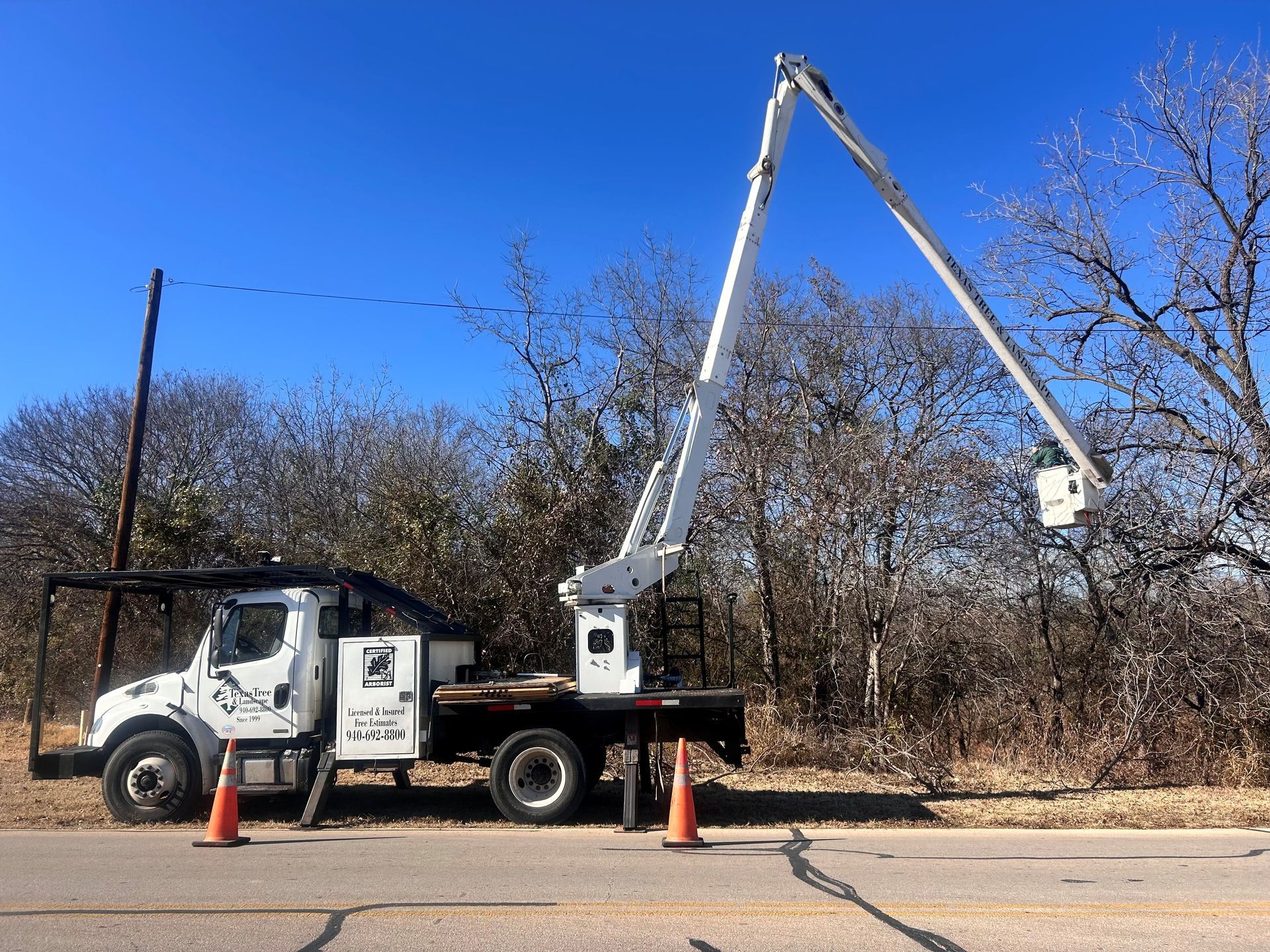 White utility truck with raised boom lift parked by roadside, with orange cones and leafless trees nearby.