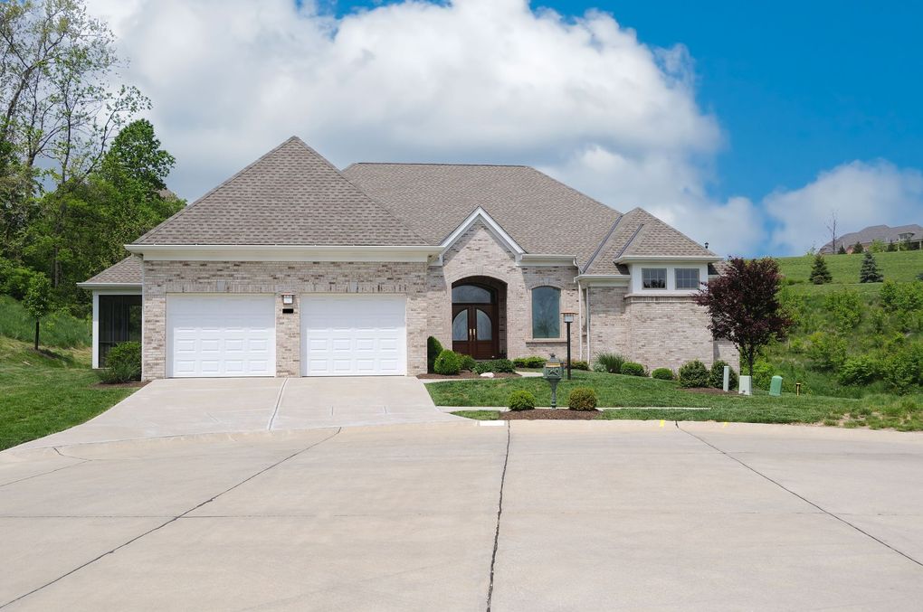 Single-story brick house with a two-car garage and a concrete driveway on a grassy, suburban hillside under a blue sky.