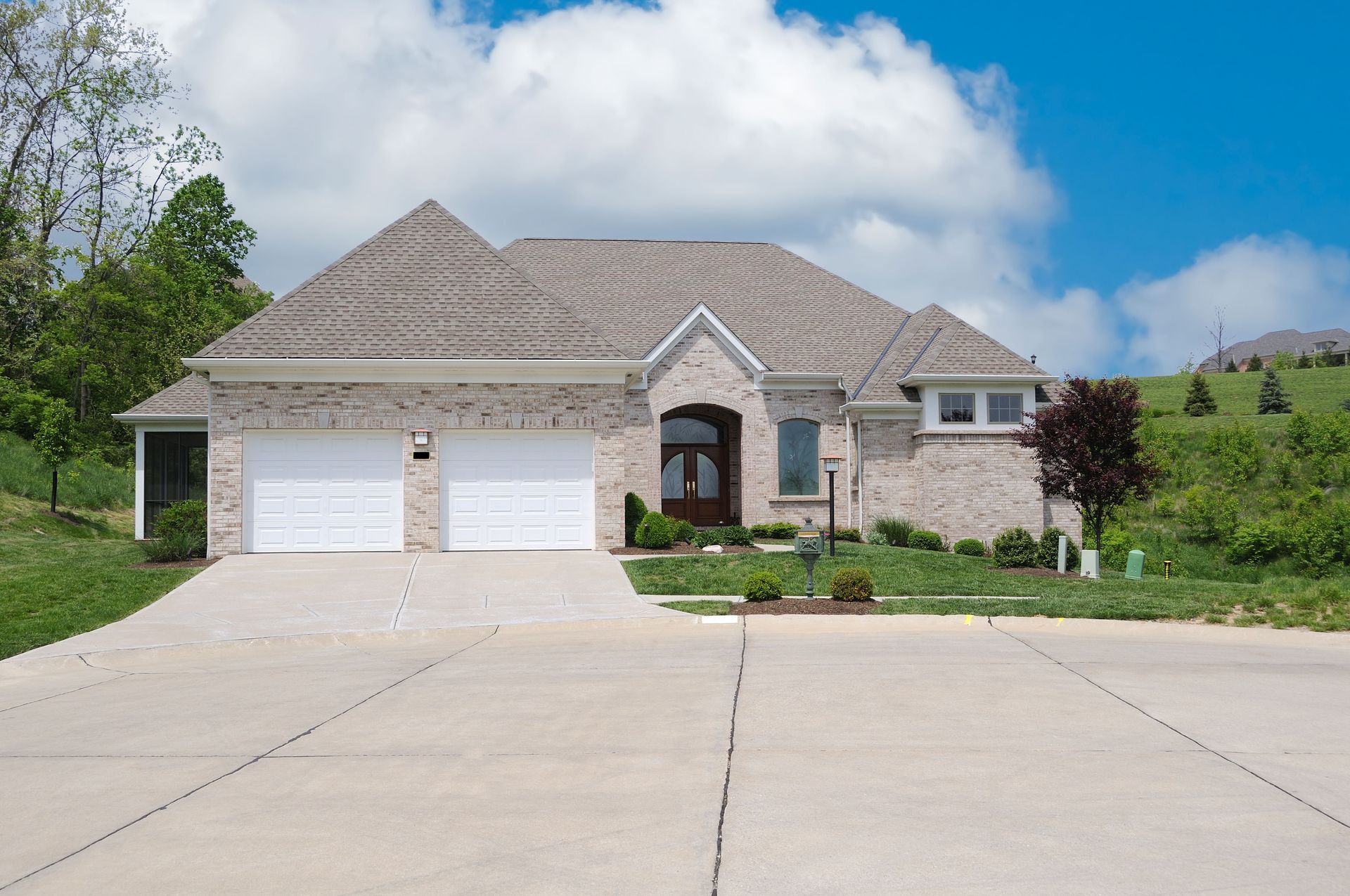 Single-story brick house with a two-car garage and a concrete driveway on a grassy, suburban hillside under a blue sky.