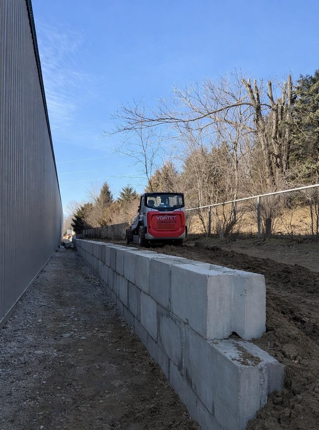 A red skid steer sits behind a low concrete block retaining wall beside a metal-sided building on a sunny day.