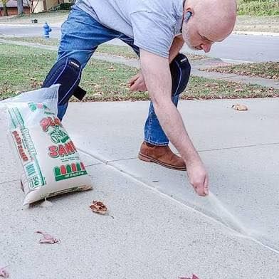 A person wearing knee pads crouches on a driveway, pouring sand from a bag into a concrete expansion joint.